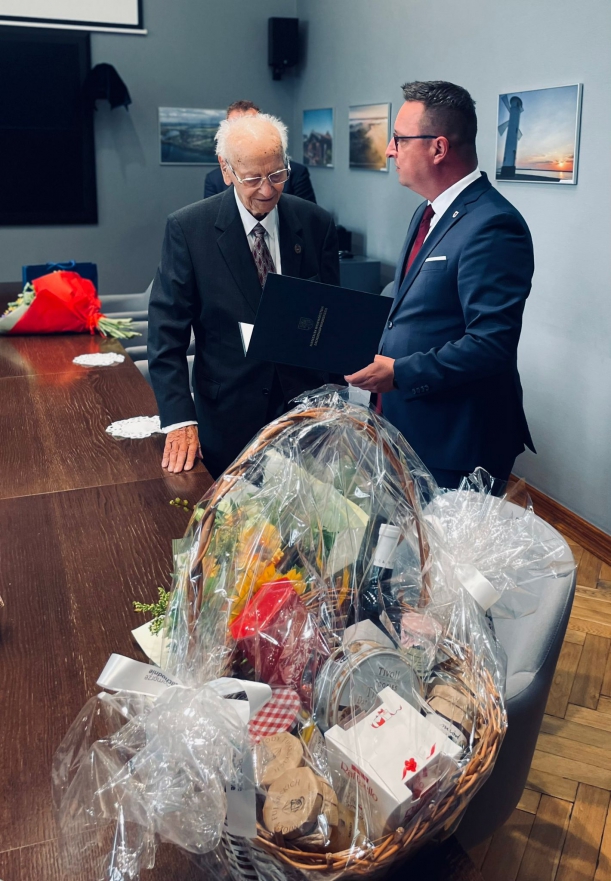 Przedstawiciel samorządu województwa składa jubilatowi życzenia, obok stół, w tle ściany pokoju.