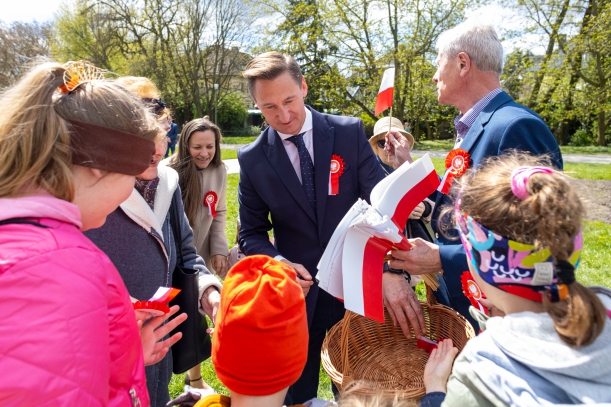 Święto Narodowe Trzeciego Maja - uroczystości przy Pomniku Czynu Polaków. Marszałek Olgierd Geblewicz rozdaje biało-czerwone chorągiewski i kotyliony. Przy nim stoi troje dzieci i kilka starszych osób.