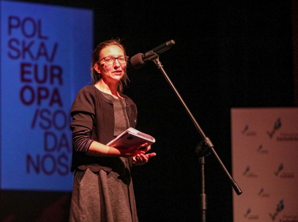 Kobieta w ciemnoszarej sukience i czarnym sweterku stoi na scenie, przy mikrofonie. W rękach trzyma książkę. W tle, na ekranie wyświetlony napis "Polska. Europa. Solidarność."