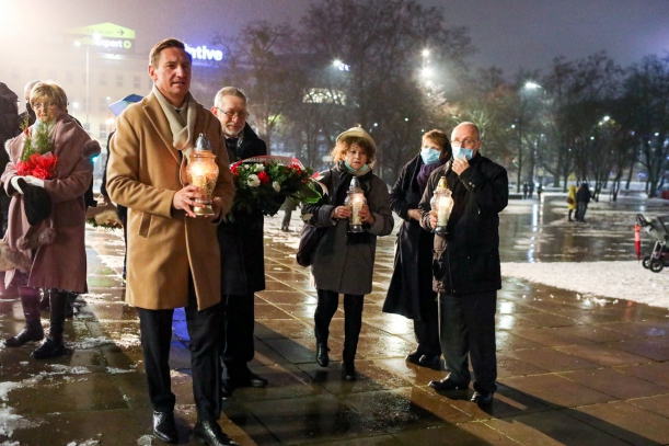 Plan Solidarności w Szczecinie, po zmroku, pada deszcz, cztery osoby (marszałek Olgierd Geblewicz, Edward Radziewicz, Franҫoise Breton–Bałuką i Jacku Challot) stoją przy pomniku Anioła Wolności, trzymają znicze i kwiaty.