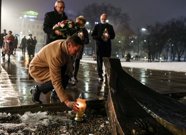 Marszałek Olgierd Geblewicz przyklęka i zapala znicz przy pomniku Anioła Wolności. Obok stoją trzy osoby, również trzymają w rękach znicze oraz wiązankę z białych i czerwonych kwiatów.