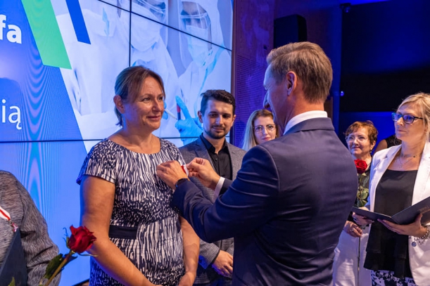 Marszałek przypina odznakę kobiecie, w tle grupa osób i baner. 