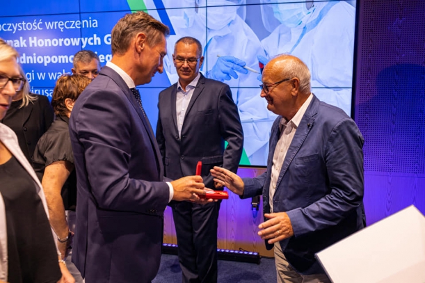 Marszałek przypina odznakę mężczyznie dyrektorowi WSPR, w tle baner.