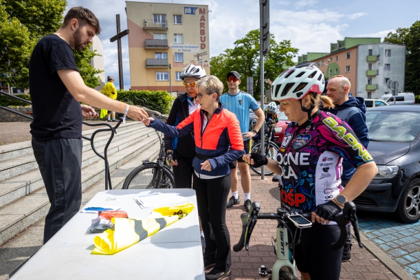 Start rajdu, uczestnicy pobierają numery startowe, w tle przestrzeń miejska. 
