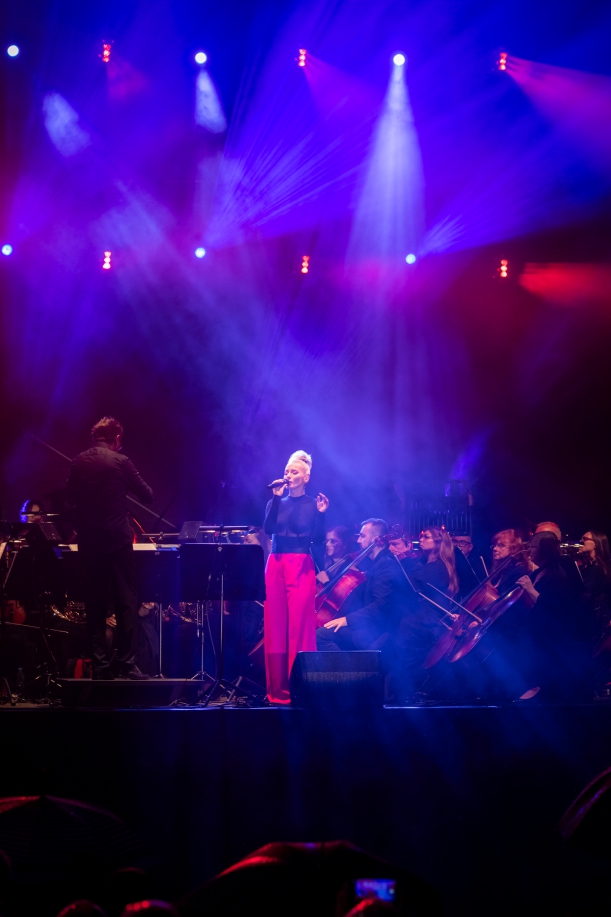 Koncert "Sierpniowe Przełomy" na placu Solidarności w Szczecinie, 27 sierpnia 2023 r. Na oświetlonej scenie - orkiestra, dyrygent i wokalistka (Sylwia Różycka), w czerwonych spodniach i czarnej bluzce.