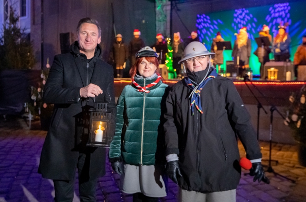Marszałek Olgierd Geblewicz na dużym dziedzińcu Zamku Książąt Pomorskich pozuje do zdjęcia z Betlejemskim Światłem Pokoju. Obok stoją dwie kobiety w harcerskich czapkach, pod kurtkami widać mundury.