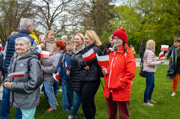Dzień Flagi Rzeczypospolitej Polskiej. Uroczystości przy Pomniku Czynu Polaków w Szczecinie. Grupa roześmianych osób, trzymających biało-czerwone chorągiewki, obserwuje uroczystości. Wysoki mężczyzna rozdaje chorągiewki.