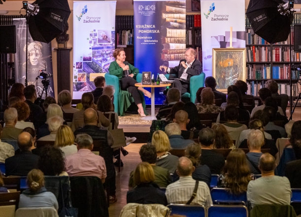 Spotkanie "Sydonia i Gryfici" (z cyklu "Otwarta społeczność") w Książnicy Pomorskiej. Na scenie - pisarka Elżbieta Cherezińska i prowadzący debatę o. Tomasz Dostatni. Na pierwszym planie - publiczność. Kilkadziesiąt osób siedzi na krzesłach.