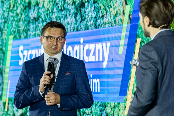 Profesor Robert Czerniawski, naukowiec z Uniwersytetu Szczecińskiego, w granatowym garniturze w kratkę stoi na scenie, trzymając przed sobą mikrofon. Obok stoi mężczyzna w garniturze. W tle jest duży ekran wyświetlający napis "Szczyt Ekologiczny".