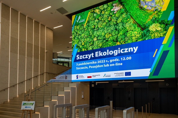 Przestronny wysoki hol Posejdon Center w Szczecinie. Po prawej stronie wielki ledowy ekran wyświetla napis "Szczyt Ekologiczny", datę, miejsce i znaki graficzne, m.in. logo Pomorza Zachdniego i funduszy UE. Po lewej stronie - szerokie schody.