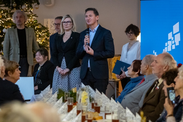 Wigilia z kombatantami w Zakładzie Aktywności Zawodowej w Dobrej. Marszałek Olgierd Geblewicz przemawia przez mikrofon. Obok niego stoi członkini zarządu województwa Anna Bańkowska. Na pierwszm planie - siedzący przy stołach seniorzy.