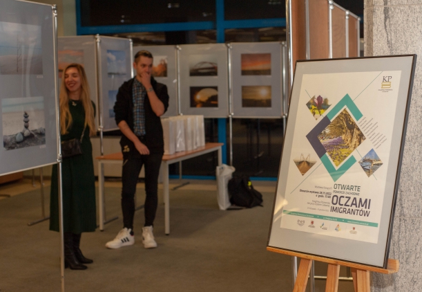 Wernisaż wystawy "Otwarte Pomorze Zachodnie. Oczami Imigrantów" w Książnicy Pomorskiej w Szczecinie. Dwie osoby oglądają fotografie wyeksponowane na tabliacach umieszczonych na stelażach. Na pierwszym planie - plakat informujący o wydarzeniu.