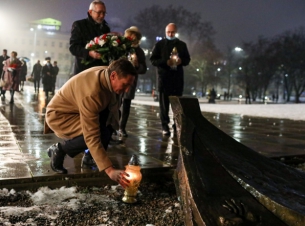 Marszałek Olgierd Geblewicz przyklęka i zapala znicz przy pomniku Anioła Wolności. Obok stoją trzy osoby, również trzymają w rękach znicze oraz wiązankę z białych i czerwonych kwiatów.
