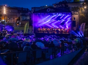 Koncert "Sierpniowe Przełomy" na placu Solidarności w Szczecinie, 27 sierpnia 2023 r. Duża plenerowa scena oświetlona na fioletowo. Na scenie - kilkudziesięciu muzyków i dyrygent. Na pierwszym planie - publiczność, ludzie stoją pod parasolami.