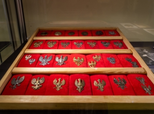 Eksponaty wystawy "Polskie orły", przypinki z wojskowych czapek i mundurów ułożone na czerwonym suknie i oświetlone w wystawowej ramie.